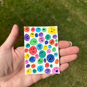 K&ouml;nnte beinhalten: Ein kleines, rechteckiges Kunstwerk mit einer Ansammlung farbenfroher, l&auml;chelnder Gesichter. Die Gesichter sind in verschiedenen Farben, darunter Rot, Blau, Gr&uuml;n, Gelb und Lila, und sind mit einfachen schwarzen Umrissen gezeichnet. Das Kunstwerk wird in einer Hand gehalten.