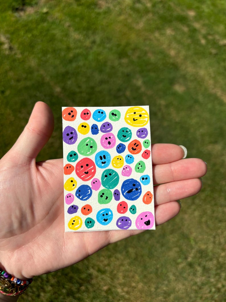 K&ouml;nnte beinhalten: Ein kleines, rechteckiges Kunstwerk mit einer Ansammlung farbenfroher, l&auml;chelnder Gesichter. Die Gesichter sind in verschiedenen Farben, darunter Rot, Blau, Gr&uuml;n, Gelb und Lila, und sind mit einfachen schwarzen Umrissen gezeichnet. Das Kunstwerk wird in einer Hand gehalten.