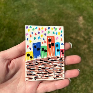 K&ouml;nnte beinhalten: Handgemalte Stadtlandschaft auf einer kleinen rechteckigen Karte. Die Geb&auml;ude sind in leuchtendem Blau, Orange, Gr&uuml;n und Gelb mit schwarzen Fenstern dargestellt. Der Himmel ist mit bunten, sternf&ouml;rmigen Konfetti gef&uuml;llt.