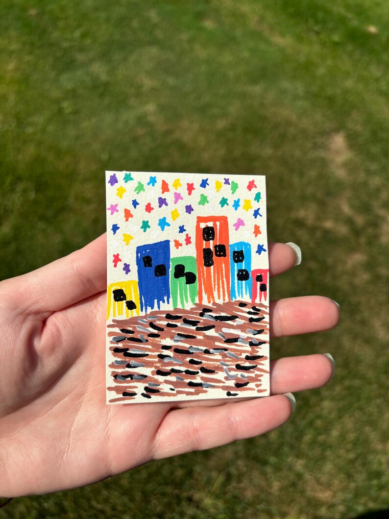 K&ouml;nnte beinhalten: Handgemalte Stadtlandschaft auf einer kleinen rechteckigen Karte. Die Geb&auml;ude sind in leuchtendem Blau, Orange, Gr&uuml;n und Gelb mit schwarzen Fenstern dargestellt. Der Himmel ist mit bunten, sternf&ouml;rmigen Konfetti gef&uuml;llt.