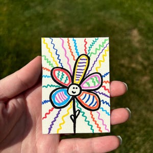 K&ouml;nnte beinhalten: Handgezeichnete Blumenillustration auf einer kleinen rechteckigen Karte. Die Blume hat ein l&auml;chelndes Gesicht und Bl&uuml;tenbl&auml;tter in verschiedenen Farben mit einem schwarzen Stiel. Der Hintergrund zeigt lebendige, wellenf&ouml;rmige Linien in mehreren Farben.