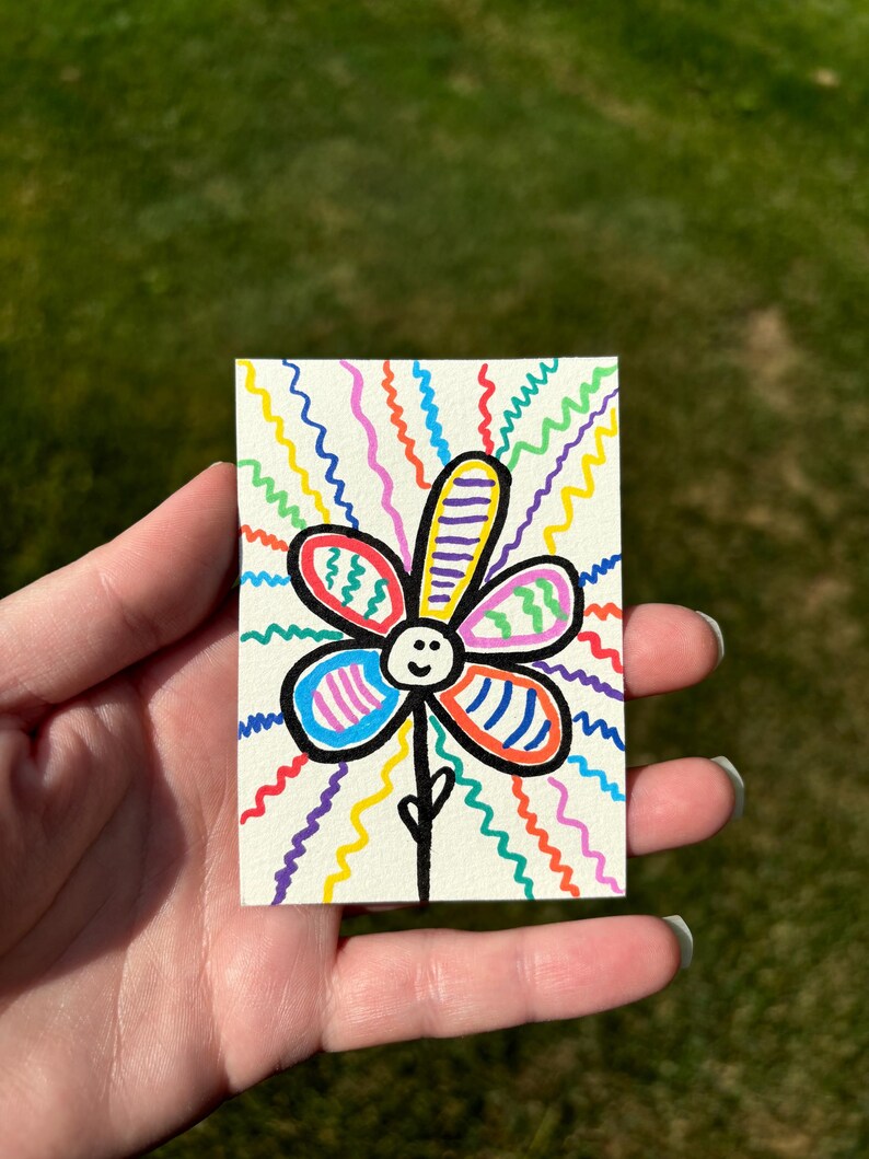 K&ouml;nnte beinhalten: Handgezeichnete Blumenillustration auf einer kleinen rechteckigen Karte. Die Blume hat ein l&auml;chelndes Gesicht und Bl&uuml;tenbl&auml;tter in verschiedenen Farben mit einem schwarzen Stiel. Der Hintergrund zeigt lebendige, wellenf&ouml;rmige Linien in mehreren Farben.