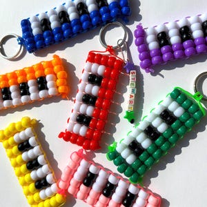 May include: A collection of colorful beaded keychains. Each keychain features a rectangular design with a pattern of black and white beads, resembling piano keys. Keychains are in various colors, including blue, orange, red, yellow, pink, purple, and green.