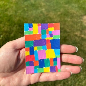 K&ouml;nnte beinhalten: Ein kleines, rechteckiges Kunstwerk mit einem Mosaik aus bunten Quadraten. Die Quadrate sind in den Farbt&ouml;nen Rot, Orange, Gelb, Gr&uuml;n, Blau, Lila und Rosa gehalten. Das Kunstwerk wird in einer Hand vor einem verschwommenen gr&uuml;nen Hintergrund gehalten.