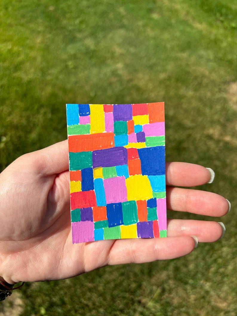 K&ouml;nnte beinhalten: Ein kleines, rechteckiges Kunstwerk mit einem Mosaik aus bunten Quadraten. Die Quadrate sind in den Farbt&ouml;nen Rot, Orange, Gelb, Gr&uuml;n, Blau, Lila und Rosa gehalten. Das Kunstwerk wird in einer Hand vor einem verschwommenen gr&uuml;nen Hintergrund gehalten.