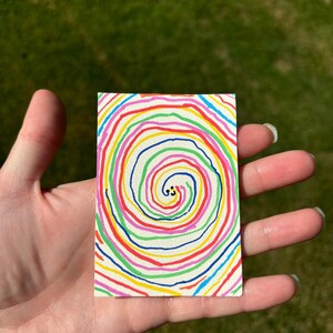 K&ouml;nnte beinhalten: Ein kleines, rechteckiges Kunstwerk mit einem farbenfrohen Spiralmuster. Die Spirale besteht aus konzentrischen Ringen in verschiedenen Farben, darunter Rot, Gelb, Gr&uuml;n und Blau. Zwei kleine schwarze Punkte befinden sich in der Mitte. Das Kunstwerk wird in einer Hand gehalten.
