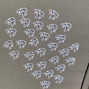 May include: A collection of small, white and black beetle stickers arranged on a textured, olive-green surface. The stickers are in a repeating pattern, with each beetle facing a different direction.
