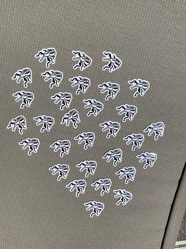 May include: A collection of small, white and black beetle stickers arranged on a textured, olive-green surface. The stickers are in a repeating pattern, with each beetle facing a different direction.