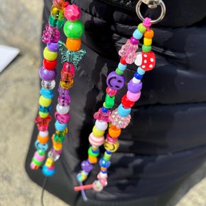 May include: Two beaded bag charms with colourful beads in various shapes and sizes. One charm features a butterfly charm, while the other has a smiley face bead and a red and white polka dot bead. The charms are attached to silver-coloured metal clasps.