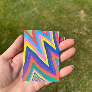 K&ouml;nnte beinhalten: Ein kleines, rechteckiges Kunstwerk mit einem lebendigen Zickzackmuster in den Farben Rot, Orange, Gelb, Gr&uuml;n, Blau und Lila. Das Kunstwerk wird in einer Hand vor einem verschwommenen gr&uuml;nen Hintergrund gehalten.