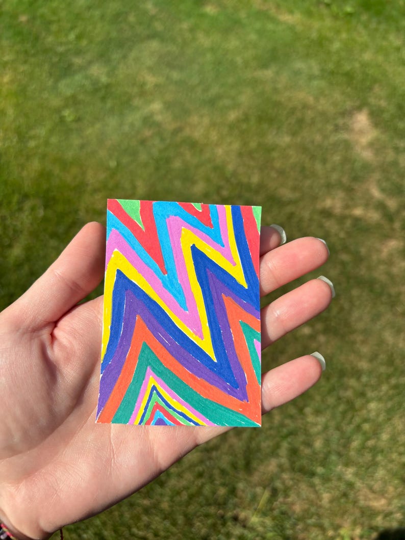 K&ouml;nnte beinhalten: Ein kleines, rechteckiges Kunstwerk mit einem lebendigen Zickzackmuster in den Farben Rot, Orange, Gelb, Gr&uuml;n, Blau und Lila. Das Kunstwerk wird in einer Hand vor einem verschwommenen gr&uuml;nen Hintergrund gehalten.