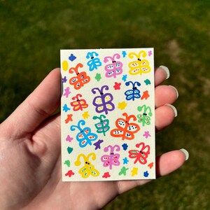 K&ouml;nnte beinhalten: Kleine, rechteckige Karte mit handgemalten Schmetterlingen in leuchtenden Farben. Die Schmetterlinge sind gelb, blau, rosa und gr&uuml;n, mit schwarzen Punktdetails. Der Hintergrund ist cremefarben, und die Karte wird in einer Hand gehalten.