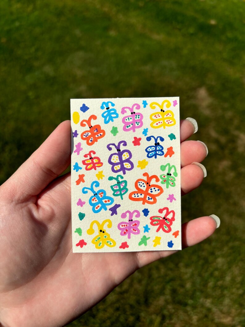 K&ouml;nnte beinhalten: Kleine, rechteckige Karte mit handgemalten Schmetterlingen in leuchtenden Farben. Die Schmetterlinge sind gelb, blau, rosa und gr&uuml;n, mit schwarzen Punktdetails. Der Hintergrund ist cremefarben, und die Karte wird in einer Hand gehalten.