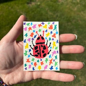 K&ouml;nnte beinhalten: Ein kleines, rechteckiges Kunstwerk mit einem roten Marienk&auml;fer mit schwarzen Punkten und Beinen. Der Marienk&auml;fer ist auf einem wei&szlig;en Hintergrund zentriert, der mit bunten sternf&ouml;rmigen Designs verziert ist. Das Kunstwerk wird in einer Hand vor einem verschwommenen gr&uuml;nen Hintergrund gehalten.
