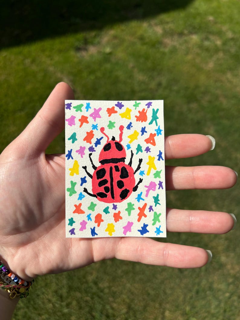 K&ouml;nnte beinhalten: Ein kleines, rechteckiges Kunstwerk mit einem roten Marienk&auml;fer mit schwarzen Punkten und Beinen. Der Marienk&auml;fer ist auf einem wei&szlig;en Hintergrund zentriert, der mit bunten sternf&ouml;rmigen Designs verziert ist. Das Kunstwerk wird in einer Hand vor einem verschwommenen gr&uuml;nen Hintergrund gehalten.