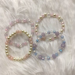 May include: Four beaded bracelets in pastel colors. The bracelets feature a mix of pearls and faceted beads in shades of pink, lavender, and clear, with some iridescent accents. The bracelets are arranged on a fluffy white surface.