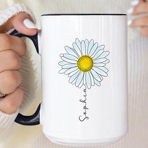 May include: White ceramic mug with a black handle and rim, featuring a daisy illustration with a yellow center and light blue petals. The name "Sophia" is written in script below the flower. The mug has the text "LesBleus" in the upper left corner.