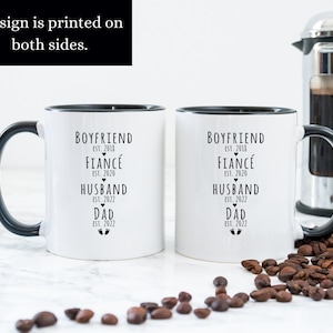 May include: Two white ceramic mugs with black handles and black rims. The mugs have black text that reads "Boyfriend Est. 2018, Fianc&eacute; Est. 2020, Husband Est. 2022, Dad Est. 2022" with a heart symbol next to "Fianc&eacute;" and a footprint symbol next to "Dad".