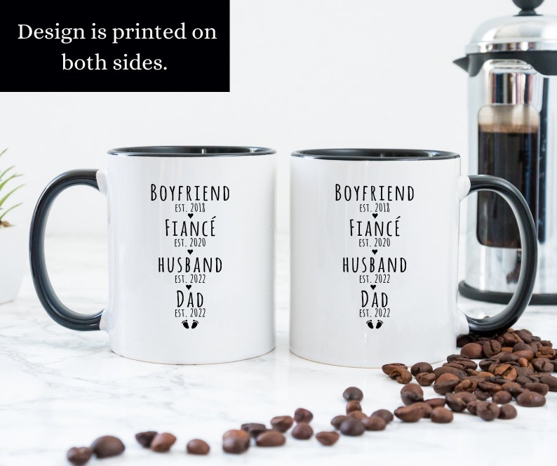 May include: Two white ceramic mugs with black handles and black rims. The mugs have black text that reads "Boyfriend Est. 2018, Fianc&eacute; Est. 2020, Husband Est. 2022, Dad Est. 2022" with a heart symbol next to "Fianc&eacute;" and a footprint symbol next to "Dad".