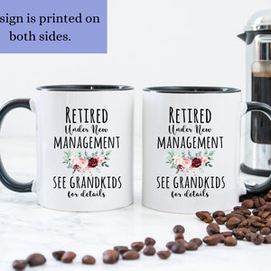 May include: Two white coffee mugs with black rims and handles. Both mugs feature the text "Retired Under New Management" with a floral design and the text "See Grandkids for details".