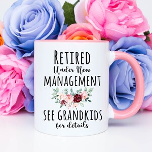 May include: White ceramic coffee mug with a pink rim and handle. The mug has black text that reads "Retired Under New Management See Grandkids for details" with a floral design.