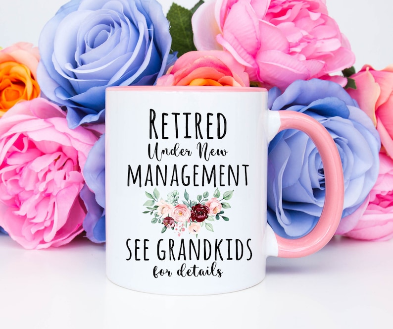 May include: White ceramic coffee mug with a pink rim and handle. The mug has black text that reads "Retired Under New Management See Grandkids for details" with a floral design.