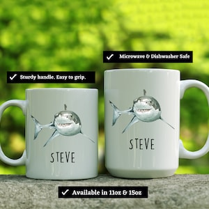 May include: Two white ceramic mugs with a shark graphic and the name "STEVE" printed on them. The mugs are available in 11oz and 15oz sizes. They are microwave and dishwasher safe.