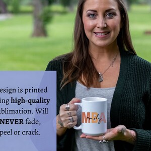 Może przedstawiać: Biały kubek ceramiczny z napisem "MBA Emily" w kolorze pomarańczowym, r&oacute;żowym i brązowym. Kubek jest trzymany przez rękę.