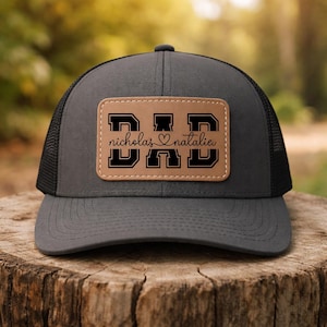 May include: A gray trucker hat with a black mesh back and a brown leather patch. The patch features the word "DAD" in bold black letters, with "nicholas" and "natalie" below, separated by a heart symbol.