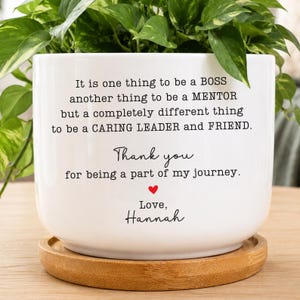 May include: A white ceramic planter with a bamboo saucer featuring black printed text that reads: It is one thing to be a BOSS another thing to be a MENTOR but a completely different thing to be a CARING LEADER and FRIEND. Thank you for being a part of my journey. Love, Hannah, with a small red heart.