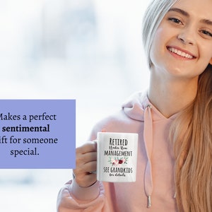 May include: White ceramic mug with black text that reads "RETIRED Under New MANAGEMENT SEE GRANDKIDS for details". The mug has a floral design with pink, red, and green flowers.