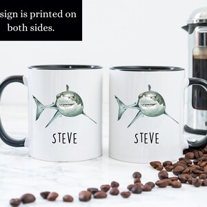 May include: Two white ceramic mugs with black handles and black rims. Each mug features a watercolor illustration of a white shark with a green and gray body. The text "STEVE" is printed in black below each shark. The mugs are sitting on a white surface with coffee beans scattered around them.