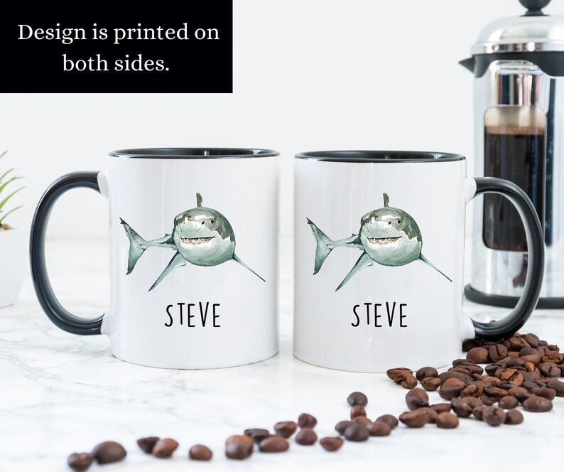 May include: Two white ceramic mugs with black handles and black rims. Each mug features a watercolor illustration of a white shark with a green and gray body. The text "STEVE" is printed in black below each shark. The mugs are sitting on a white surface with coffee beans scattered around them.
