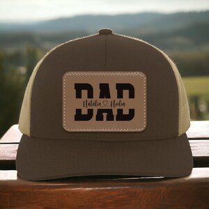 K&ouml;nnte beinhalten: Braune und beigefarbene Trucker-Kappe mit einem braunen Leder-Patch, auf dem "DAD" steht, darunter die Namen "Natalia" und "Noella".