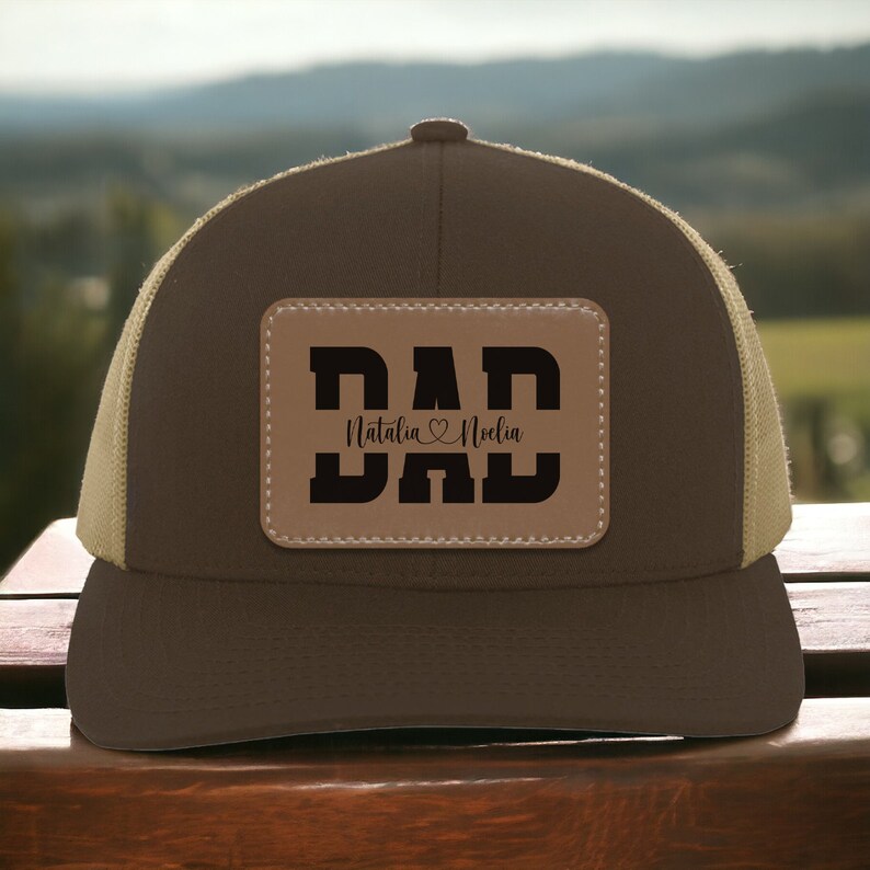 K&ouml;nnte beinhalten: Braune und beigefarbene Trucker-Kappe mit einem braunen Leder-Patch, auf dem "DAD" steht, darunter die Namen "Natalia" und "Noella".