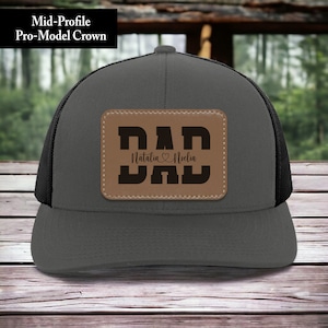 K&ouml;nnte beinhalten: Eine graue und schwarze Trucker-Kappe mit einem braunen Leder-Patch, auf dem "DAD" steht, darunter die Namen "Natalia" und "Noelin".