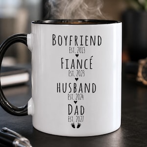 May include: A white ceramic mug with a black handle and interior rim. The front features black text reading: BOYFRIEND EST. 2013, FIANCÉ EST. 2023, HUSBAND EST. 2024, DAD EST. 2027, separated by small heart icons and a pair of baby footprints at the bottom.