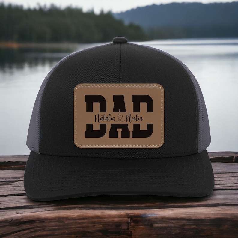 K&ouml;nnte beinhalten: Schwarze und graue Trucker-Kappe mit einem braunen Leder-Patch, auf dem "DAD" steht, darunter die Namen "Natalia" und "Noelle".