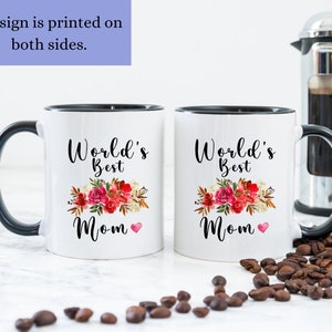 May include: Two white ceramic mugs with black handles and black rims. Both mugs feature a floral design with the text "World's Best Mom" and a small red heart. The mugs are on a white surface with coffee beans scattered around them.