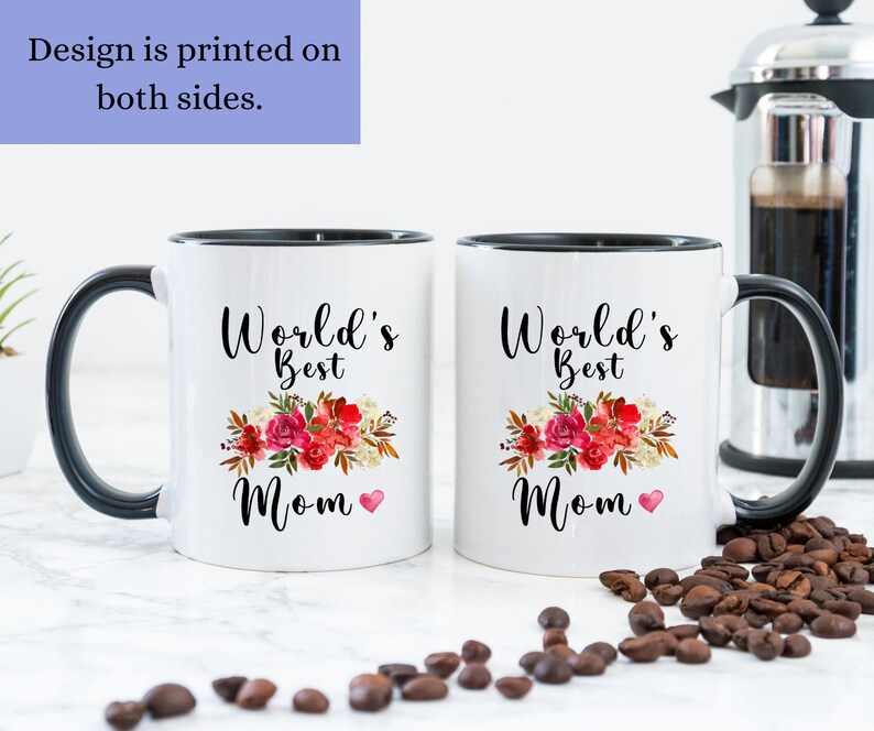 May include: Two white ceramic mugs with black handles and black rims. Both mugs feature a floral design with the text "World's Best Mom" and a small red heart. The mugs are on a white surface with coffee beans scattered around them.