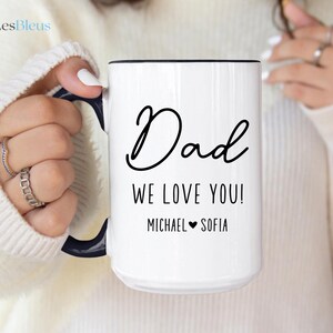 May include: White ceramic mug with a black handle and rim. The mug features the word "Dad" in a cursive font, along with the message "WE LOVE YOU!" and the names "MICHAEL" and "SOFIA" with a heart symbol.