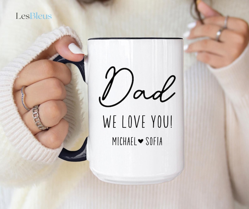 May include: White ceramic mug with a black handle and rim. The mug features the word "Dad" in a cursive font, along with the message "WE LOVE YOU!" and the names "MICHAEL" and "SOFIA" with a heart symbol.