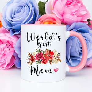 May include: White ceramic mug with a pink rim and handle. The mug has a floral design and the text "World's Best Mom" with a small heart.