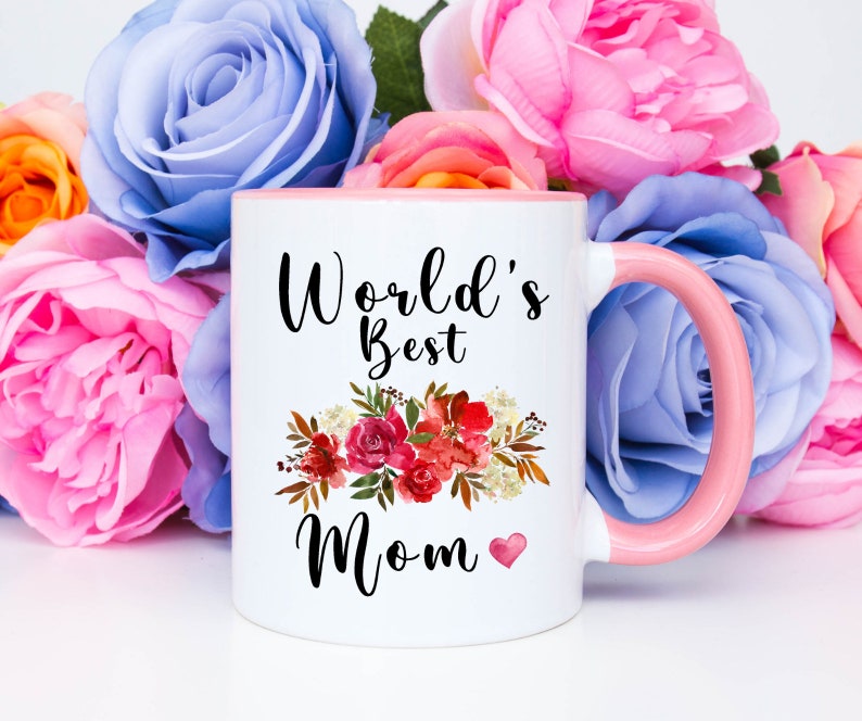 May include: White ceramic mug with a pink rim and handle. The mug has a floral design and the text "World's Best Mom" with a small heart.