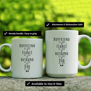 May include: Two white ceramic mugs with black text. The text on the mugs reads "Boyfriend Est. 2018, Fianc&eacute; Est. 2020, Husband Est. 2022, Dad Est. 2022". The mugs are microwave and dishwasher safe and available in 11oz and 15oz sizes.