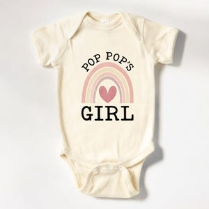 May include: A cream-colored baby onesie with a rainbow and heart design. The text "Pop Pop's Girl" is printed on the onesie.