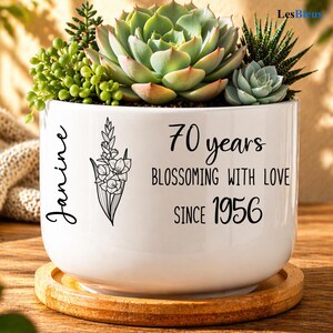 May include: White ceramic planter with a variety of succulents. The planter has the text "Janine", "70 years, Blossoming with Love, Since 1956" and a floral illustration. The planter sits on a wooden coaster.