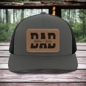 K&ouml;nnte beinhalten: Eine graue und schwarze Trucker-Kappe mit einem braunen Leder-Patch, auf dem "DAD" steht, darunter die Namen "Natalia" und "Noelin".