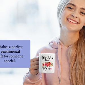 May include: White ceramic mug with a floral design and the text "World's Best Mom" in black lettering. The mug is held by a person's hand.