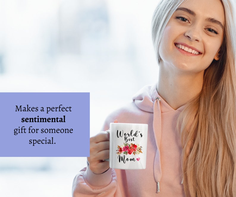 May include: White ceramic mug with a floral design and the text "World's Best Mom" in black lettering. The mug is held by a person's hand.